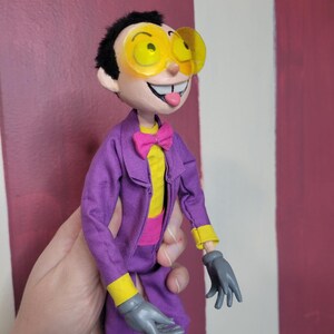 May include: A handmade doll of a cartoon character with black hair, yellow-tinted glasses, and a purple suit. The doll has a yellow shirt, pink bow tie, and gray gloves. The doll's tongue is sticking out.