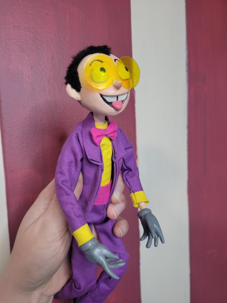 May include: A handmade doll of a cartoon character with black hair, yellow-tinted glasses, and a purple suit. The doll has a yellow shirt, pink bow tie, and gray gloves. The doll's tongue is sticking out.