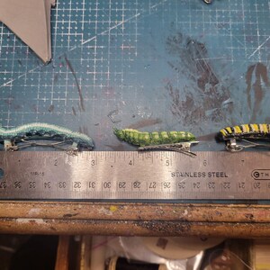 K&ouml;nnte beinhalten: Drei bunte Haarklammern in Form von Raupen. Die Klammern sind aus Stoff und haben eine metallische Oberfl&auml;che. Die Klammern sind auf einem Lineal mit den Zahlen 1 bis 9 angeordnet.