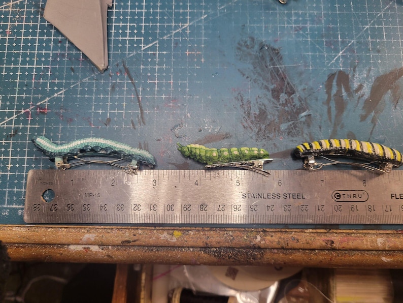 K&ouml;nnte beinhalten: Drei bunte Haarklammern in Form von Raupen. Die Klammern sind aus Stoff und haben eine metallische Oberfl&auml;che. Die Klammern sind auf einem Lineal mit den Zahlen 1 bis 9 angeordnet.