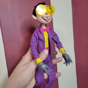 May include: A handmade doll dressed in a purple suit with yellow accents, gray gloves, and yellow-tinted glasses. The doll has a pink bow tie and a yellow shirt. The doll is smiling with its tongue sticking out.