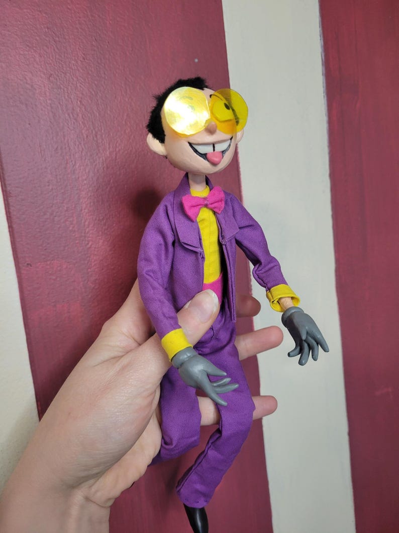 May include: A handmade doll dressed in a purple suit with yellow accents, gray gloves, and yellow-tinted glasses. The doll has a pink bow tie and a yellow shirt. The doll is smiling with its tongue sticking out.