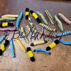 K&ouml;nnte beinhalten: Eine Sammlung von 15 bunten Raupen-Haarklammern. Die Klammern sind aus Filz und haben eine Vielzahl von Farben, darunter Schwarz, Gelb, Gr&uuml;n, Blau und Pink. Die Klammern sind alle in verschiedenen Gr&ouml;&szlig;en und Formen, und einige haben schwarze und wei&szlig;e Punkte.