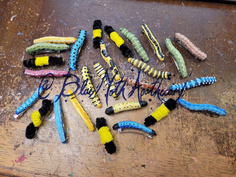 K&ouml;nnte beinhalten: Eine Sammlung von 15 bunten Raupen-Haarklammern. Die Klammern sind aus Filz und haben eine Vielzahl von Farben, darunter Schwarz, Gelb, Gr&uuml;n, Blau und Pink. Die Klammern sind alle in verschiedenen Gr&ouml;&szlig;en und Formen, und einige haben schwarze und wei&szlig;e Punkte.