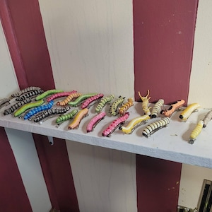 K&ouml;nnte beinhalten: Eine Sammlung von 20 bunten Raupen-Haarklammern. Die Klammern sind aus Kunststoff und haben eine Vielzahl von Farben, darunter Pink, Gr&uuml;n, Gelb und Schwarz. Die Klammern sind in einer Reihe auf einem wei&szlig;en Regal angeordnet.