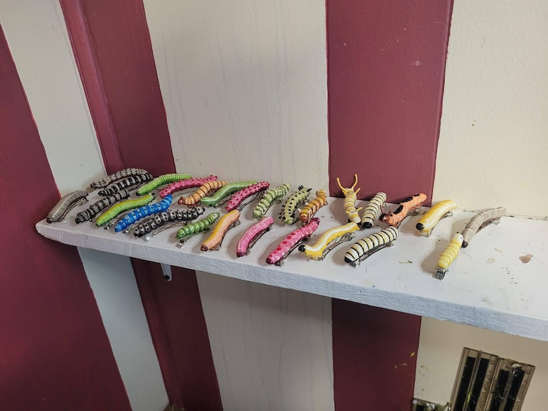 K&ouml;nnte beinhalten: Eine Sammlung von 20 bunten Raupen-Haarklammern. Die Klammern sind aus Kunststoff und haben eine Vielzahl von Farben, darunter Pink, Gr&uuml;n, Gelb und Schwarz. Die Klammern sind in einer Reihe auf einem wei&szlig;en Regal angeordnet.