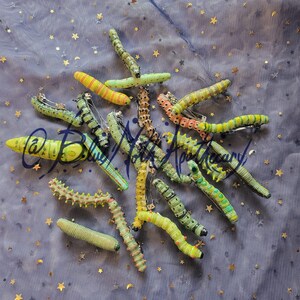 K&ouml;nnte beinhalten: Eine Sammlung von 15 handbemalten Raupen-Haarklammern in verschiedenen Gr&uuml;nt&ouml;nen, Gelb und Orange. Die Klammern sind auf einem blauen Hintergrund mit goldenen Sternen angeordnet.