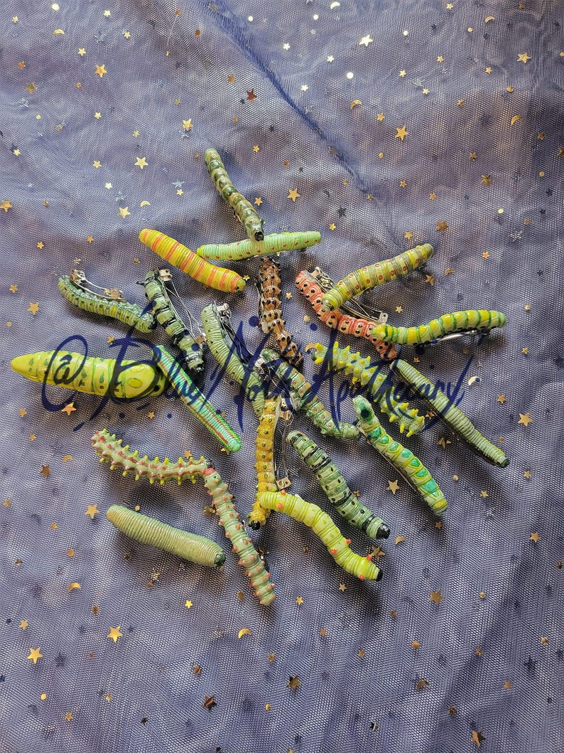 K&ouml;nnte beinhalten: Eine Sammlung von 15 handbemalten Raupen-Haarklammern in verschiedenen Gr&uuml;nt&ouml;nen, Gelb und Orange. Die Klammern sind auf einem blauen Hintergrund mit goldenen Sternen angeordnet.