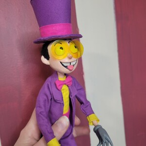 May include: A handmade doll of a cartoon character wearing a purple suit, top hat, and yellow-tinted glasses. The doll has a yellow shirt, pink bow tie, and gray gloves. The doll's tongue is sticking out.