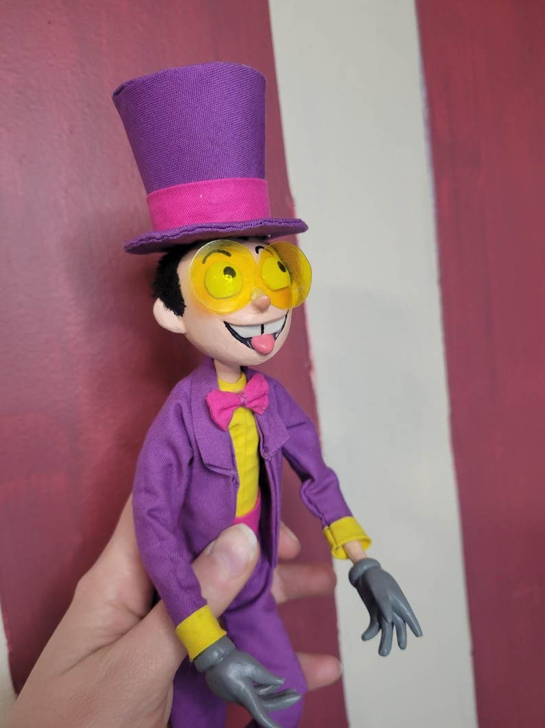 May include: A handmade doll of a cartoon character wearing a purple suit, top hat, and yellow-tinted glasses. The doll has a yellow shirt, pink bow tie, and gray gloves. The doll's tongue is sticking out.