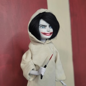 May include: A doll with black hair, blue eyes, and a wide red smile, wearing a white hooded robe. The doll holds a small knife with a red blade. The background has red and white stripes.