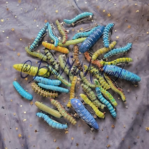 K&ouml;nnte beinhalten: Eine Sammlung von handbemalten, bunten Raupen-Haarklammern. Die Klammern sind aus Polymer Clay gefertigt und in verschiedenen Blaut&ouml;nen, Gr&uuml;nt&ouml;nen und Gelbt&ouml;nen gehalten. Jede Klammer hat ein einzigartiges Design und ist perfekt, um jeder Frisur einen Hauch von Verspieltheit zu verleihen.