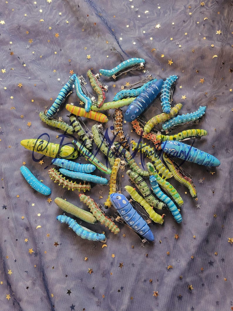 K&ouml;nnte beinhalten: Eine Sammlung von handbemalten, bunten Raupen-Haarklammern. Die Klammern sind aus Polymer Clay gefertigt und in verschiedenen Blaut&ouml;nen, Gr&uuml;nt&ouml;nen und Gelbt&ouml;nen gehalten. Jede Klammer hat ein einzigartiges Design und ist perfekt, um jeder Frisur einen Hauch von Verspieltheit zu verleihen.