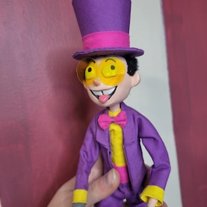 May include: A handmade doll dressed in a purple suit, yellow shirt, and pink bow tie. The doll has a purple top hat with a pink band and yellow-tinted glasses. The doll's gloves are gray, and it has a smiling expression with its tongue sticking out.