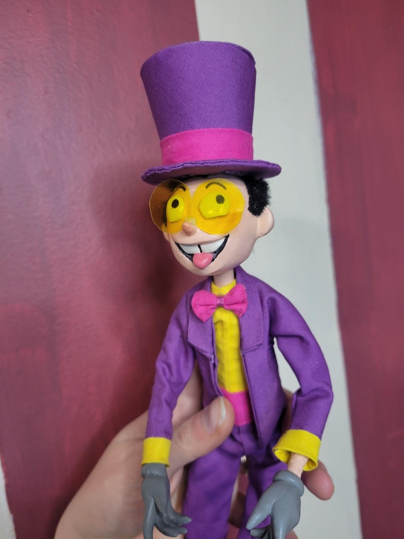 May include: A handmade doll dressed in a purple suit, yellow shirt, and pink bow tie. The doll has a purple top hat with a pink band and yellow-tinted glasses. The doll's gloves are gray, and it has a smiling expression with its tongue sticking out.