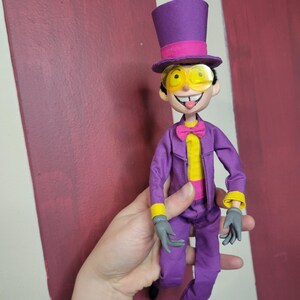 May include: A doll dressed in a purple suit, top hat, and yellow-tinted glasses. The doll has a yellow shirt, pink bow tie, and gray gloves. The background is a maroon and white striped wall.
