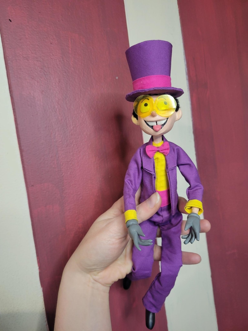 May include: A doll dressed in a purple suit, top hat, and yellow-tinted glasses. The doll has a yellow shirt, pink bow tie, and gray gloves. The background is a maroon and white striped wall.