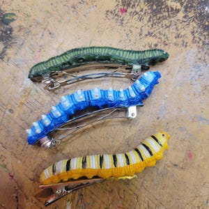 K&ouml;nnte beinhalten: Vier Haarklammern in Form von Raupen in verschiedenen Farben. Die Klammern sind gr&uuml;n, blau, gelb und schwarz-wei&szlig;.