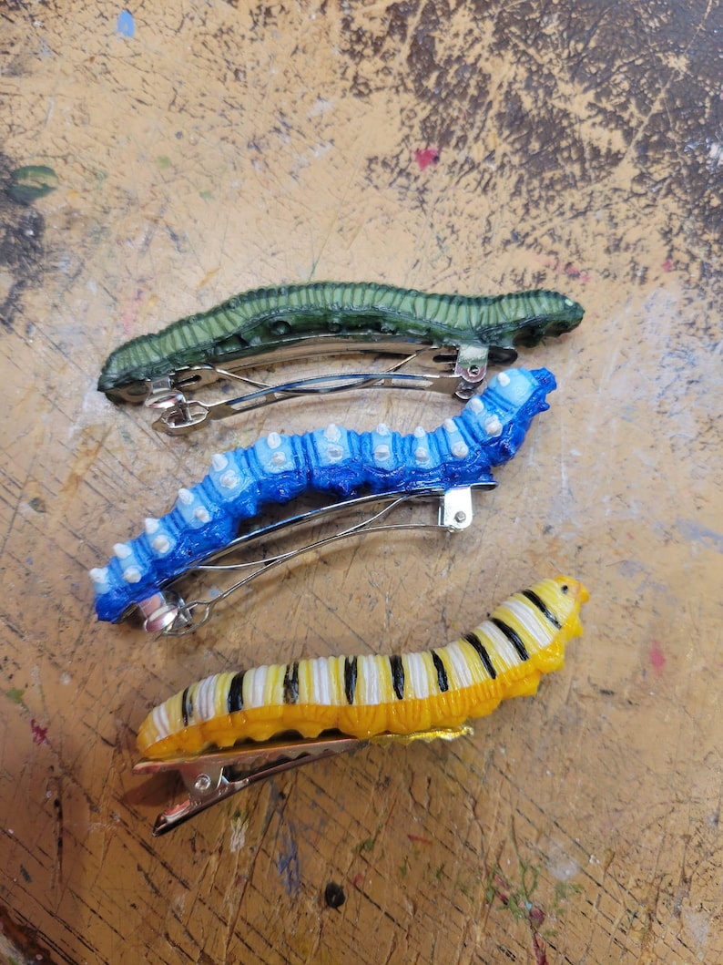 K&ouml;nnte beinhalten: Vier Haarklammern in Form von Raupen in verschiedenen Farben. Die Klammern sind gr&uuml;n, blau, gelb und schwarz-wei&szlig;.