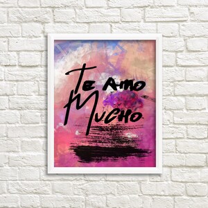 Op de afbeelding: Een wit ingelijst afdruk met een roze, blauwe en oranje aquarel achtergrond. De woorden "Te Amo Mucho" zijn in zwarte verf op de afdruk geschreven.