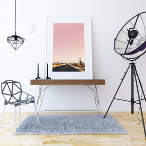 May include: A white framed photograph of a pink sky with a road leading to a tree in the distance. The photograph is on a wooden desk with a black metal chair and a black tripod lamp with a large reflector.