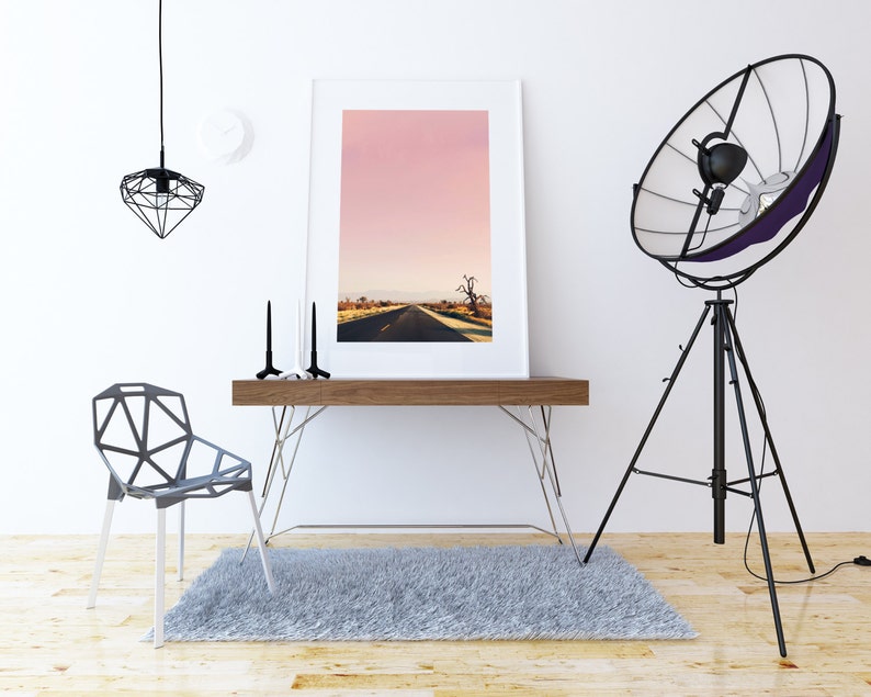 May include: A white framed photograph of a pink sky with a road leading to a tree in the distance. The photograph is on a wooden desk with a black metal chair and a black tripod lamp with a large reflector.