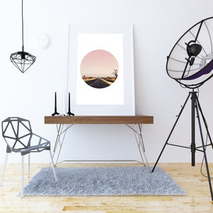 Puede incluir: Una fotograf&iacute;a enmarcada en blanco de un camino del desierto con un cielo rosa en el fondo. La foto est&aacute; colgada en una pared blanca sobre un escritorio de madera con una silla de metal negro y una l&aacute;mpara de tr&iacute;pode negra.