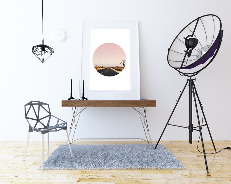 Puede incluir: Una fotograf&iacute;a enmarcada en blanco de un camino del desierto con un cielo rosa en el fondo. La foto est&aacute; colgada en una pared blanca sobre un escritorio de madera con una silla de metal negro y una l&aacute;mpara de tr&iacute;pode negra.