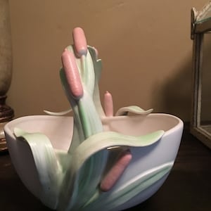 May include: A white ceramic bowl with a green and pink floral design. The bowl has a handle made of sculpted leaves and flowers.