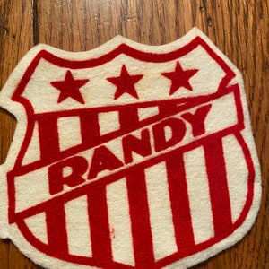 May include: Red and white felt patch with a shield shape. The shield has three stars at the top and the word "RANDY" in a banner across the middle. The shield is divided into vertical stripes.