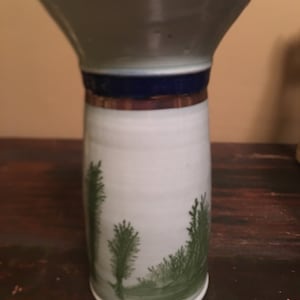 May include: A white ceramic vase with a blue and gold rim. The vase has a green painted design of trees on the front.