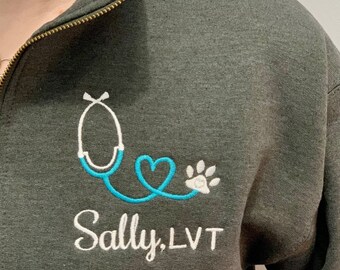 May include: A dark gray zip-up sweatshirt with a teal blue stethoscope, heart, and paw print embroidered on the left chest. The text "Sally, LVT" is embroidered below the design.