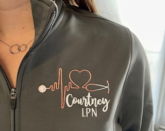 May include: A dark grey zip-up jacket with a white embroidered design featuring a heart, a stethoscope, and a heartbeat line. The text "Courtney LPN" is embroidered below the design.