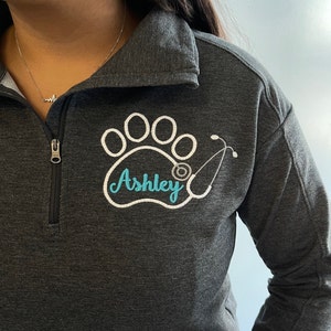 May include: A dark gray fleece pullover with a white paw print and stethoscope design. The name "Ashley" is embroidered in blue inside the paw print.