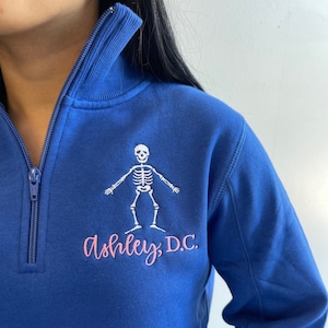 May include: A royal blue quarter-zip sweatshirt with a white skeleton graphic and pink embroidered text that reads "Ashley, D.C."