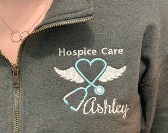 May include: A dark gray sweatshirt with a white embroidered design. The design features a stethoscope, a heart, and wings. The text "Hospice Care" and "Ashley" is also embroidered on the sweatshirt.