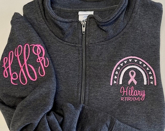 May include: A dark gray zippered sweatshirt with a pink ribbon and rainbow design on the front. The text "Hilary RTR(M)" is embroidered below the design. The left sleeve has a pink monogram "HFF".