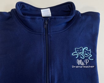 May include: A navy blue 1/4 zip sweatshirt with a light blue embroidered design of two theatre masks and the text "Ms. P Drama Teacher".