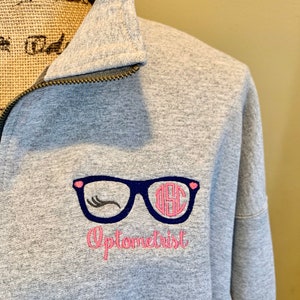 May include: A gray sweatshirt with a zipper closure. The sweatshirt has an embroidered design of a pair of glasses with eyelashes and a monogram in the lenses. The text "Optometrist" is embroidered below the glasses.