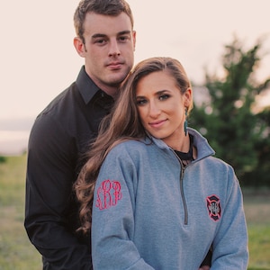 May include: A couple poses for a photo, the woman wearing a gray sweatshirt with a red monogram and a firefighter patch that says "Wife".