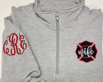 Może przedstawiać: Szara bluza z czerwono-czarną odznaką strażaka z wyszytym napisem "wife". Bluza ma zamek błyskawiczny i monogram na lewym rękawie.