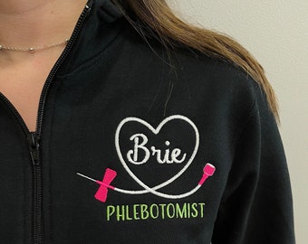 May include: Black sweatshirt with a white heart and the name "Brie" inside. The heart is connected to a white medical tubing with a pink needle. The text "PHLEBOTOMIST" is embroidered in green below the heart.