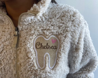 May include: A beige sherpa fleece pullover with a white tooth graphic and the name "Chelsea" embroidered on it. The tooth has a small pink heart on it.
