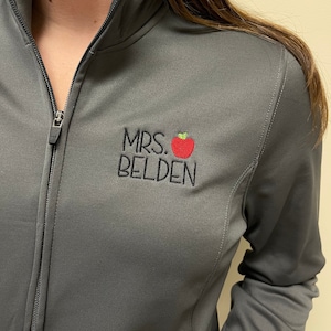May include: A gray zip-up jacket with a black embroidered design that says "MRS. BELDEN" with a red apple.
