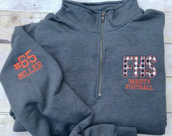 May include: A charcoal gray quarter-zip sweatshirt with orange embroidered text and a plaid patch. The left sleeve displays "#65 MILLER" in orange. The right chest features "FHS VARSITY FOOTBALL" in orange, with the letters "FHS" in a black and white plaid pattern.