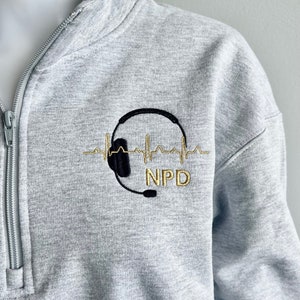 May include: A gray sweatshirt with a black and gold embroidered design. The design features a headset with a microphone and a heartbeat line. The text "NPD" is embroidered below the headset.