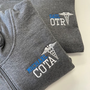 May include: Two gray zippered sweatshirts with embroidered names and the medical symbol. The top sweatshirt has "SAUL OTR" embroidered on the right side. The bottom sweatshirt has "MICHAEL COTA" embroidered on the left side.