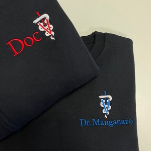 May include: Two black sweatshirts with embroidered logos. The top sweatshirt has a red embroidered logo that says "Doc" with a medical symbol. The bottom sweatshirt has a blue embroidered logo that says "Dr. Manganaro" with a medical symbol.