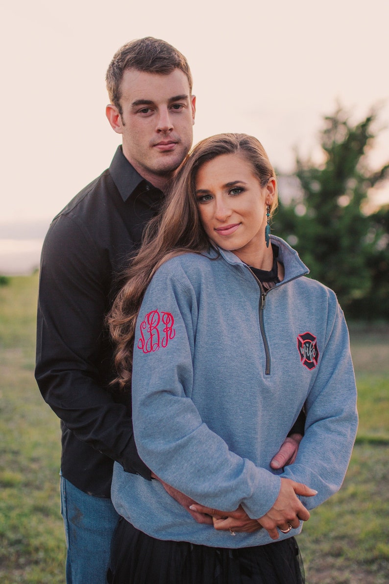 May include: A couple poses for a photo, the woman wearing a gray sweatshirt with a red monogram and a firefighter patch that says "Wife".