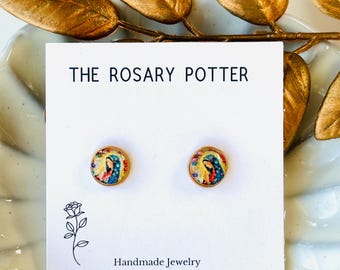 Puede incluir: Un par de pendientes de botón dorados con una imagen colorida de la Virgen María en cada pendiente. Los pendientes se muestran en una tarjeta blanca con el texto "THE ROSARY POTTER" y "Handmade Jewelry".